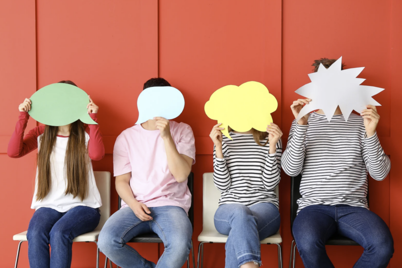 4 Junge Menschen auf einer Bank, die Pappschilder mit Sprechblasen über ihr Gesicht halten.