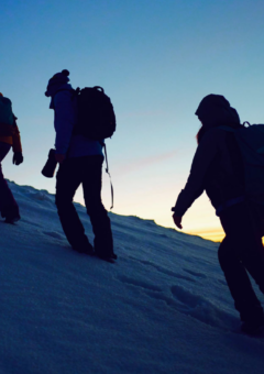 3 Menschen beim winterlichen Bergsteigen. Gehen hintereinander einen steilen Berg hinauf. Die Sonne geht im Hintergrund auf.