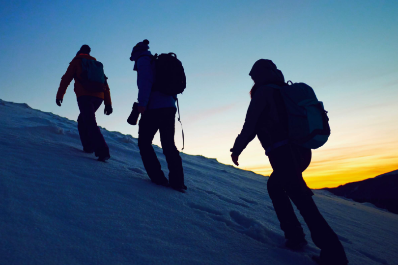 3 Menschen beim winterlichen Bergsteigen. Gehen hintereinander einen steilen Berg hinauf. Die Sonne geht im Hintergrund auf.