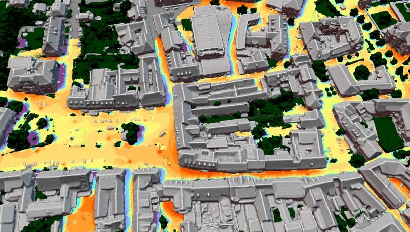 Eine Stadtansicht aus der Vogelperspektive durch eine spezielle Wärmebildkamera, die Hitzeinseln im urbanen Raum aufzeigt. Man sieht ausgegraute Gebäude und Straßen, die von gelb (kühler) bis orange (wärmer) eingefärbt sind.