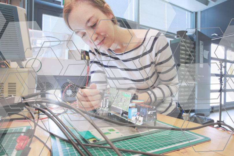 Eine Frau in einem Labor arbeitet konzentriert an einer Leiterplatte, umgeben von technischen Geräten und Kabeln. Über das Bild sind hexagonale Muster als Overlay gelegt, die Innovation und Technologie symbolisieren.