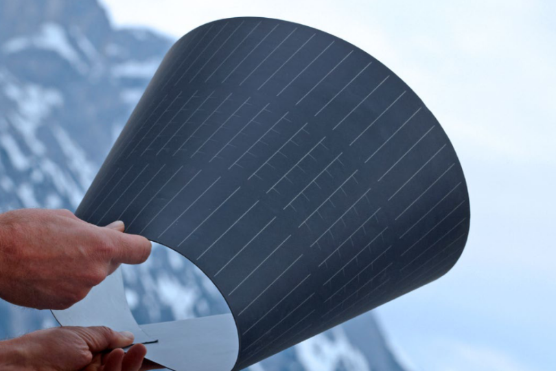 Eine flexible, gebogene Solarzelle wird von zwei Händen gehalten, mit einem schneebedeckten Berg im Hintergrund. Die dunkelblaue Oberfläche der Solarzelle zeigt feine Linien, die die einzelnen Solarzellenbereiche definieren.