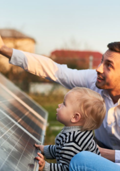 Auf dem Bild sind ein Mann, eine Frau, und ein Kind zu sehen. Der vater zeigt mit dem Finger auf einen PV-Modul. Im Hintergrund siehr man Einfamillienhäuser.