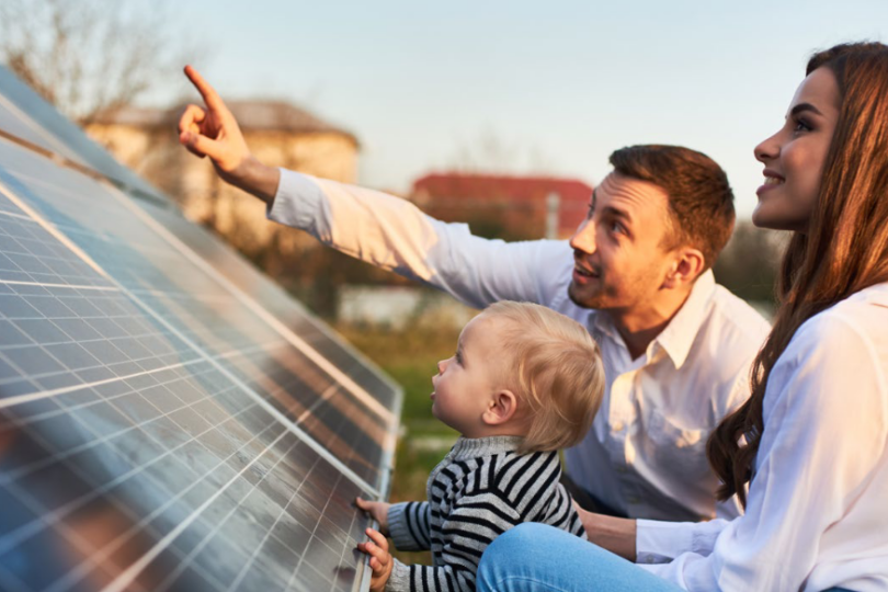 Eine Familie, bestehend aus einem Mann, einer Frau und einem kleinen Kind, sitzt neben einer Solaranlage im Freien. Der Mann zeigt lächelnd auf etwas in der Ferne, während das Kind neugierig auf die Solarmodule schaut. Die Szene vermittelt einen Eindruck von Nachhaltigkeit und Zukunftsdenken.