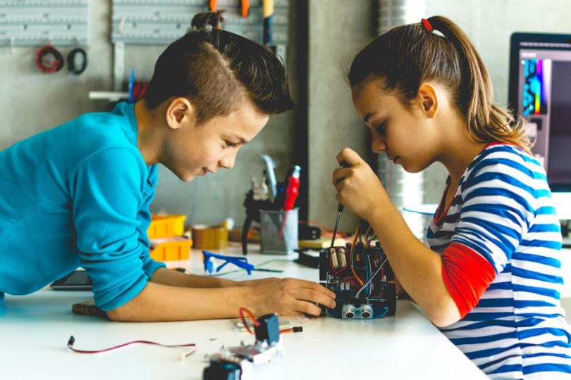 Zwei Kinder arbeiten konzentriert an einem kleinen Roboterprojekt auf einem weißen Tisch in einer Werkstatt. Der Junge im blauen Pullover und das Mädchen im gestreiften Shirt verwenden Werkzeuge, während im Hintergrund technische Geräte und Materialien zu sehen sind.