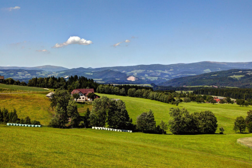 Eine idyllische Landschaft mit grünen Wiesen, vereinzelten Bäumen und einem Bauernhaus, umgeben von sanften Hügeln. Im Hintergrund erstrecken sich bewaldete Berge unter einem klaren, blauen Himmel mit wenigen Wolken.