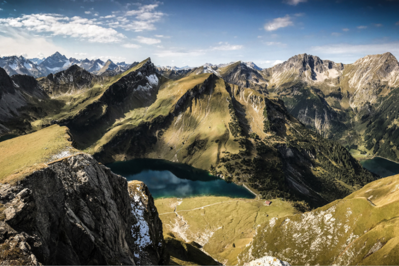 Panoramablick auf eine beeindruckende Berglandschaft mit schroffen Gipfeln, grünen Hängen und zwei klaren Bergseen. Die Szenerie ist von einem blauen Himmel mit vereinzelten Wolken gekrönt und vermittelt eine ruhige, majestätische Atmosphäre.