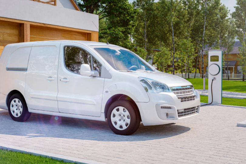 Ein weißer elektrischer Lieferwagen, der vor einer modernen Garage parkt und an einer Ladestation aufgeladen wird. Im Hintergrund sind grüne Bäume und ein gepflegter Garten zu sehen, die eine nachhaltige und umweltfreundliche Atmosphäre unterstreichen.