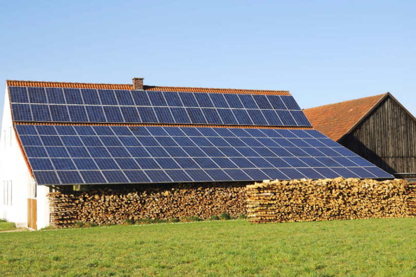 Auf dem Bild ist ein Bauernhaus zu sehen, welches eine große PV-Anlage auf dem Dach hat. Die PV-Dachanlage dient auch als Überdeckung für Brennholz.