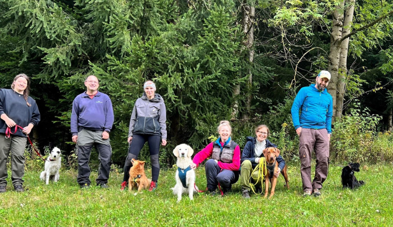 Das Bild zeigt Menschen mit ihren Hunden vor einem Wald. Die Hunde werden ausgebildet, um Borkenkäfer aufzuspüren.