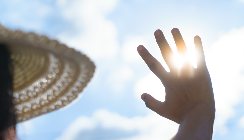 Eine Person mit Hut vor freiem Himmel blickt in Richtung Sonne und versucht sich mit der Hand vor der Sonne abzuschirmen.