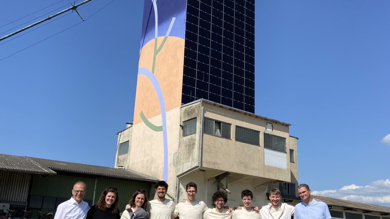 Das Bild zeigt Bernd Vogl, den Geschäftsführer des Klima- und Energiefonds, zusammen mit dem Team des Projektes „Solar Silo“. Im Hintergrund ist der Solar Silo Turm zu sehen, der teilweise mit einer Solar-Paneel-Fassade und einer abstrakten, farbenfrohen Wandgestaltung versehen ist. Die Gruppe steht bei sonnigem Wetter vor dem Turm und lächelt in die Kamera, um die erfolgreiche Eröffnung dieses innovativen Projekts zu feiern.