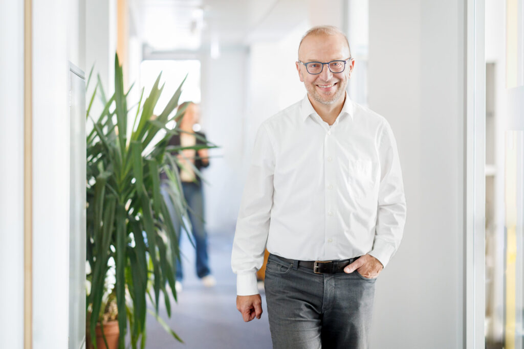 Geschäftsführer Bernd Vogl im Gang im Büro. Ein lächelnder Mann mittleren Alters mit Brille, weißem Hemd und dunkler Hose geht einen hellen Büroflur entlang. Links im Vordergrund steht eine große Grünpflanze, während im Hintergrund eine unscharfe Person zu sehen ist. Die Szene wirkt freundlich und professionell.