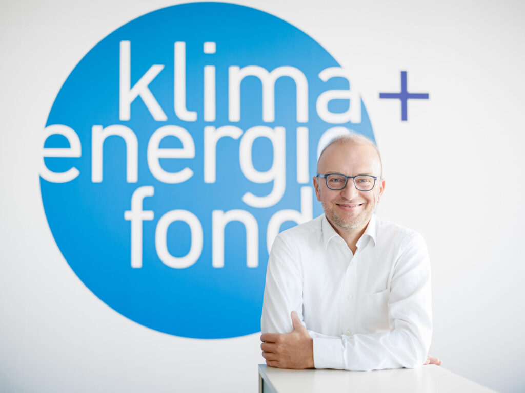 Geschäftsführer Bernd Vogl vor dem Logo des Klima- und Energiefonds. Ein Mann mit Brille, kurzem Bart und einem freundlichen Lächeln lehnt sich mit verschränkten Armen auf einen weißen Tisch. Er trägt ein weißes Hemd und steht vor einer Wand mit dem Logo von 