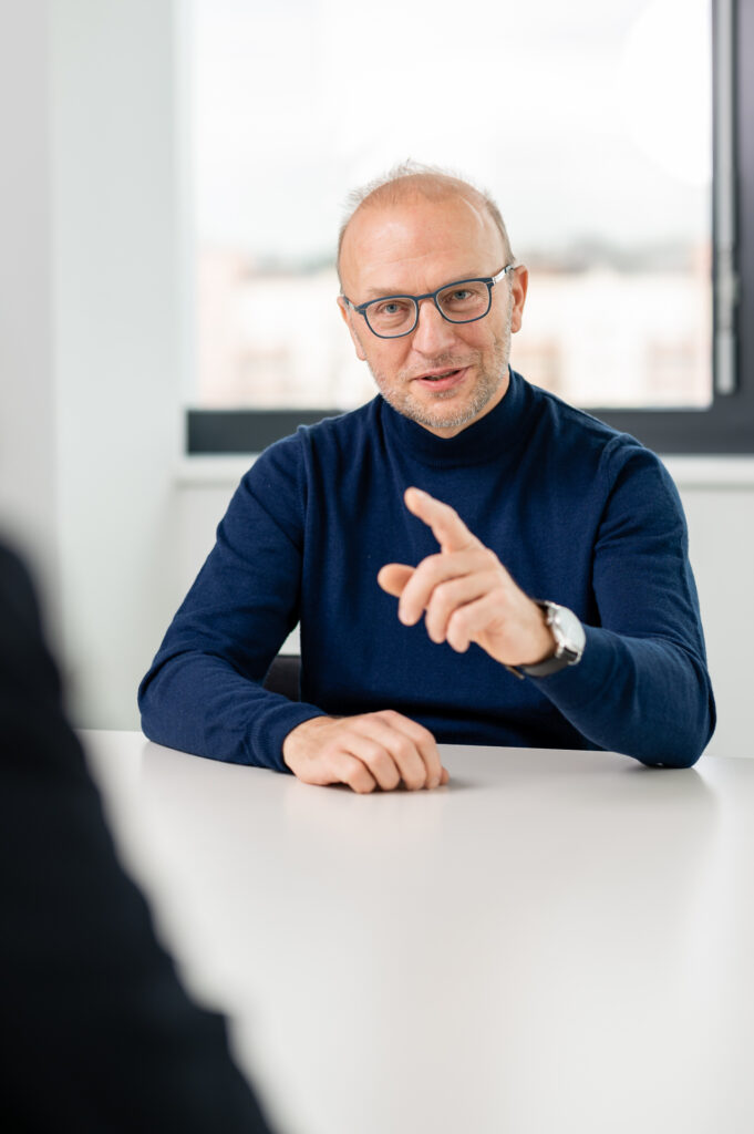 Geschäftsführer Bernd Vogl erklärt etwas am Tisch sitzend. Ein Mann mittleren Alters mit Brille und dunklem Rollkragenpullover sitzt an einem weißen Tisch und zeigt mit dem Finger in Richtung einer unscharfen Person im Vordergrund. Sein Gesichtsausdruck wirkt konzentriert und engagiert, während im Hintergrund ein großes Fenster mit Blick auf eine verschwommene Stadtlandschaft zu sehen ist.