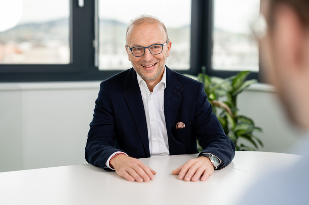 Geschäftsführer Bernd Vogl lächelnd am Tisch sitzend. Ein Mann mittleren Alters mit Brille, kurzem Bart und einem freundlichen Lächeln sitzt an einem weißen Tisch. Er trägt ein dunkelblaues Sakko mit einem weißen Hemd und einem gemusterten Einstecktuch. Im Hintergrund befindet sich ein großes Fenster mit Blick auf eine hügelige Landschaft sowie eine grüne Pflanze. Im Vordergrund ist unscharf der Kopf einer weiteren Person zu erkennen.