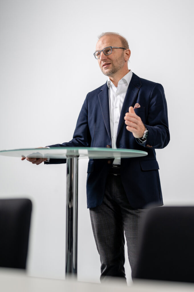 Geschäftsführer Bernd Vogl am Podium. Ein Mann mittleren Alters mit Brille und hellem Teint steht an einem runden Glastisch und hält eine Präsentation. Er trägt ein dunkelblaues Sakko, ein weißes Hemd und dunkle Hosen. Seine Gestik deutet auf eine engagierte Rede hin, während der Hintergrund schlicht und weiß gehalten ist, was den Fokus auf den Sprecher lenkt.