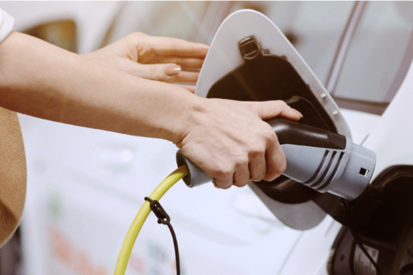 Das Bild zeigt eine Frau, die gerade ein Elektroauto auflädt. Ihre Hände halten den Ladestecker, der an einem gelben Kabel befestigt ist, und stecken ihn in die Ladebuchse des Fahrzeugs. Der Fokus liegt auf den Händen und dem Stecker, während das Auto und die Umgebung unscharf im Hintergrund zu sehen sind.