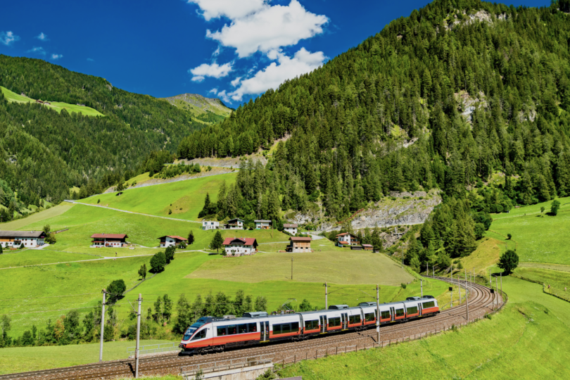 Man sieht eine österreichische Zuggarnitur der ÖBB durch eine österreichische Berg- und Hügellandschaft fahren.