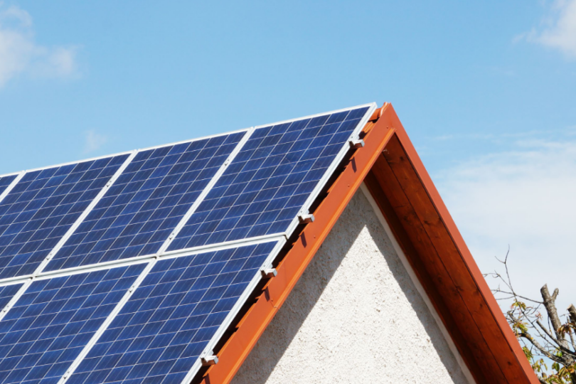 Solarmodule auf dem Schrägdach eines weißen Hauses mit Holzbalken, die in die Sonne ragen. Der blaue Himmel im Hintergrund deutet auf einen sonnigen, klaren Tag hin, während ein Baumast am rechten Bildrand sichtbar ist.