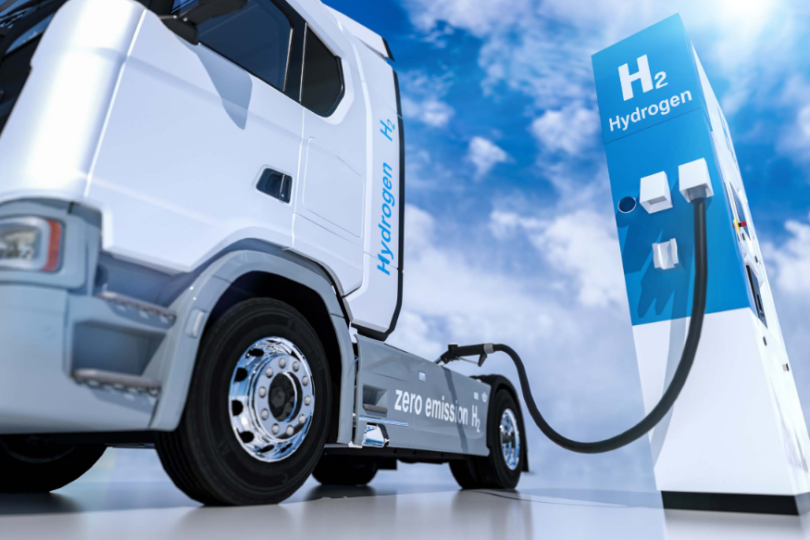 Ein weißer Wasserstoff-LKW mit der Aufschrift 'zero emission H₂' wird an einer Wasserstoff-Tankstelle betankt. Die Zapfsäule ist blau-weiß und mit 'H₂ Hydrogen' beschriftet, vor einem sonnigen Himmel mit weißen Wolken.