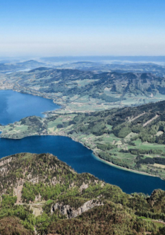 Das Bild zeigt eine Luftaufnahme eines großen Sees, eingebettet in eine hügelige Landschaft mit grünen Wiesen, Wäldern und Bergen im Hintergrund. Der See hat eine tiefblaue Farbe und eine unregelmäßige Form mit mehreren Buchten und Landzungen. Es könnte sich um einen See im Alpenvorland handeln.