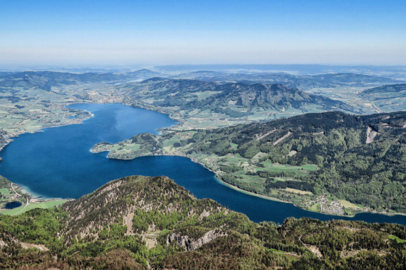 Das Bild zeigt eine Luftaufnahme eines großen Sees, eingebettet in eine hügelige Landschaft mit grünen Wiesen, Wäldern und Bergen im Hintergrund. Der See hat eine tiefblaue Farbe und eine unregelmäßige Form mit mehreren Buchten und Landzungen. Es könnte sich um einen See im Alpenvorland handeln.