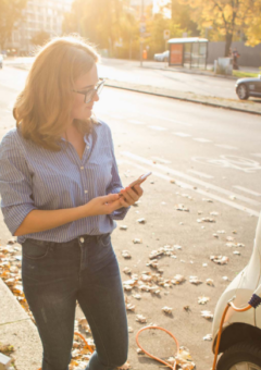 Auf dem Bild ist eine Frau zu sehen, die ein E-Fahrzeug aufladet. Im hintergrund ist eine urbane Landschaft zu sehen.