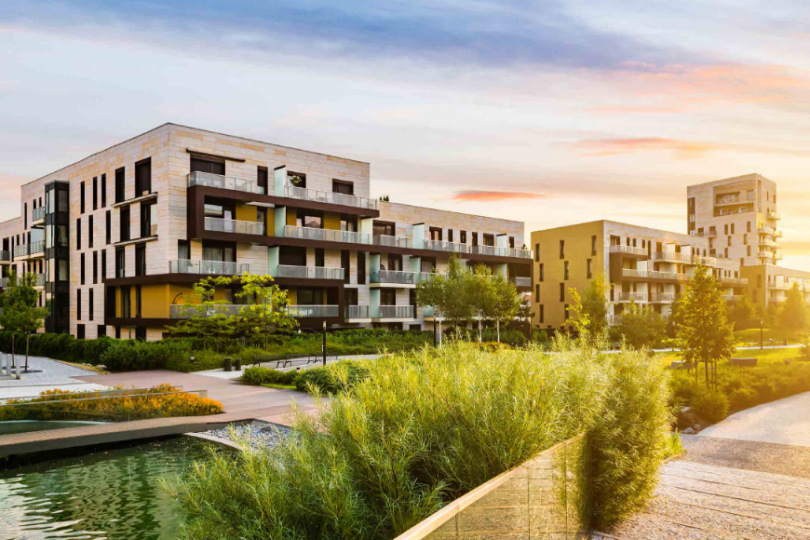 Das Bild zeigt einen modernen Wohnkomplex mit mehreren Gebäuden in verschiedenen Höhen und Farben, eingebettet in eine grüne Landschaft mit Bäumen, Sträuchern und einem kleinen Teich. Die warme Abendsonne taucht die Szene in ein goldenes Licht und erzeugt eine ruhige und einladende Atmosphäre.