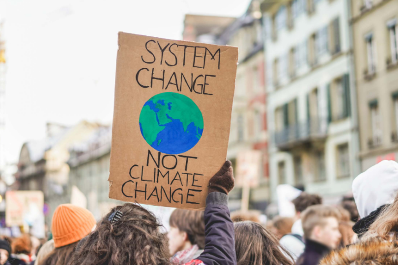 Das Bild zeigt ein Schild mit der Aufschrift "SYSTEM CHANGE NOT CLIMATE CHANGE". Der Text ist in Großbuchstaben geschrieben und in zwei Zeilen angeordnet, wobei "SYSTEM CHANGE" oben und "NOT CLIMATE CHANGE" unten steht. Das Schild scheint aus Pappe oder einem ähnlichen Material zu bestehen und wird von einer Person gehalten, deren Hand am unteren Rand des Schildes sichtbar ist. Der Hintergrund ist unscharf, aber es scheint sich um eine Außenumgebung mit Bäumen und möglicherweise anderen Personen zu handeln.