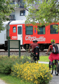 Zwei Radfahrer fahren auf dem Fahrradweg vor einem ÖBB-Zug im Hintergrund.