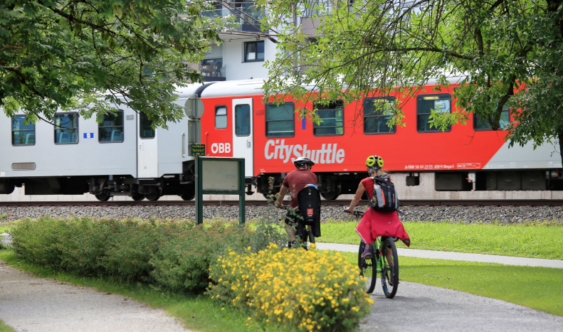 Zwei Radfahrer fahren auf dem Fahrradweg vor einem ÖBB-Zug im Hintergrund.