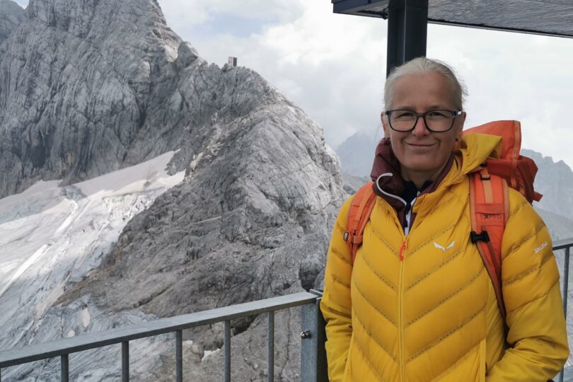 Eine Person in gelber Jacke steht auf einer Aussichtsplattform in den Bergen mit verschneiten Gipfeln im Hintergrund.