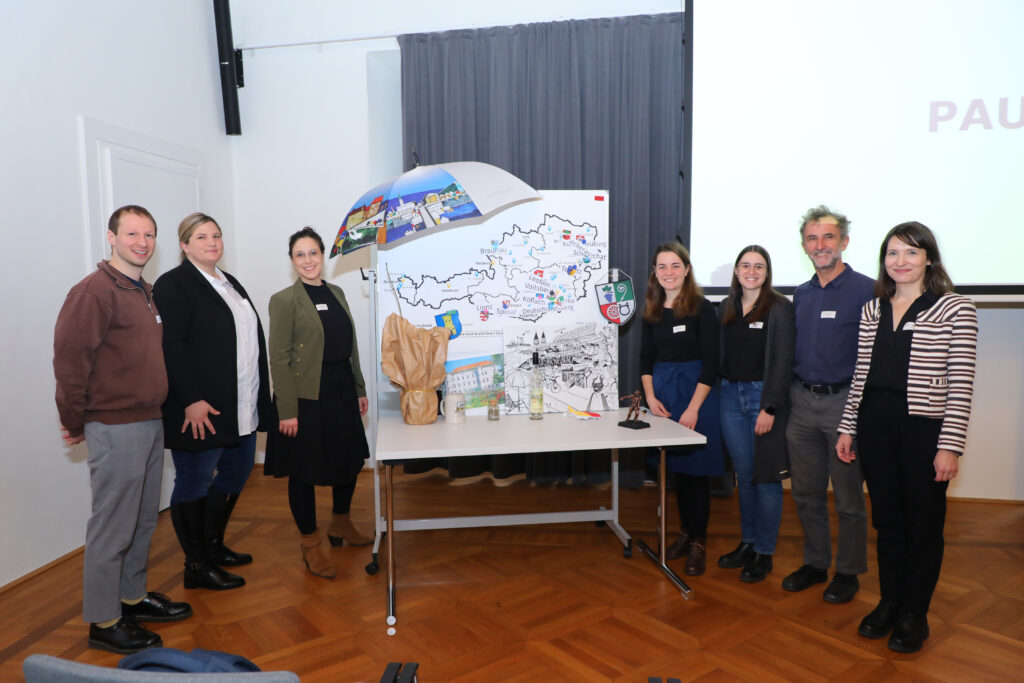 Eine Gruppe von sechs Personen steht vor einem Tisch mit einem farbenfrohen Regenschirm und einer Landkarte, auf der die lokale Kultur und Sehenswürdigkeiten dargestellt sind.