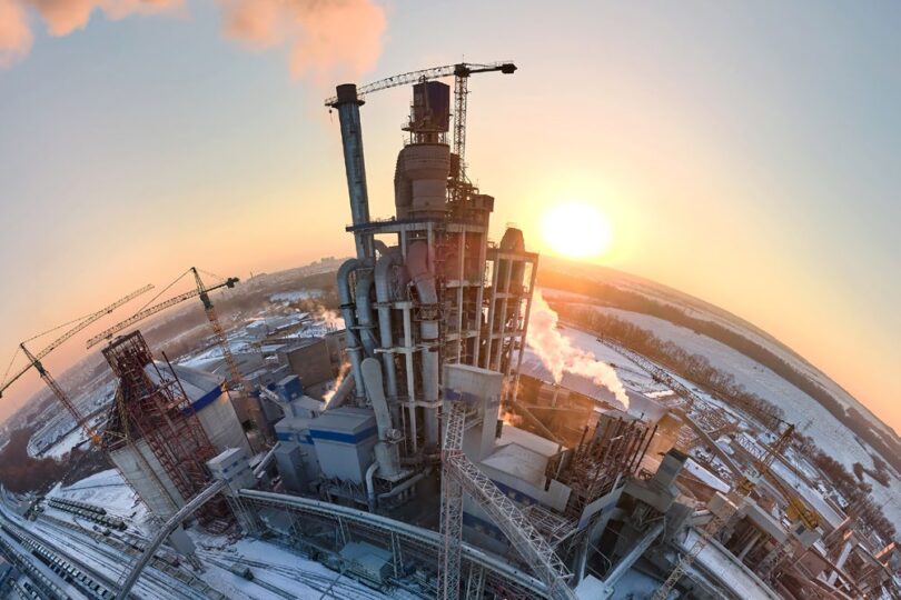 Luftaufnahme eines Industriekomplexes bei Sonnenaufgang, mit Rauch aus Schornsteinen und schneebedecktem Boden, Kräne im Vordergrund.